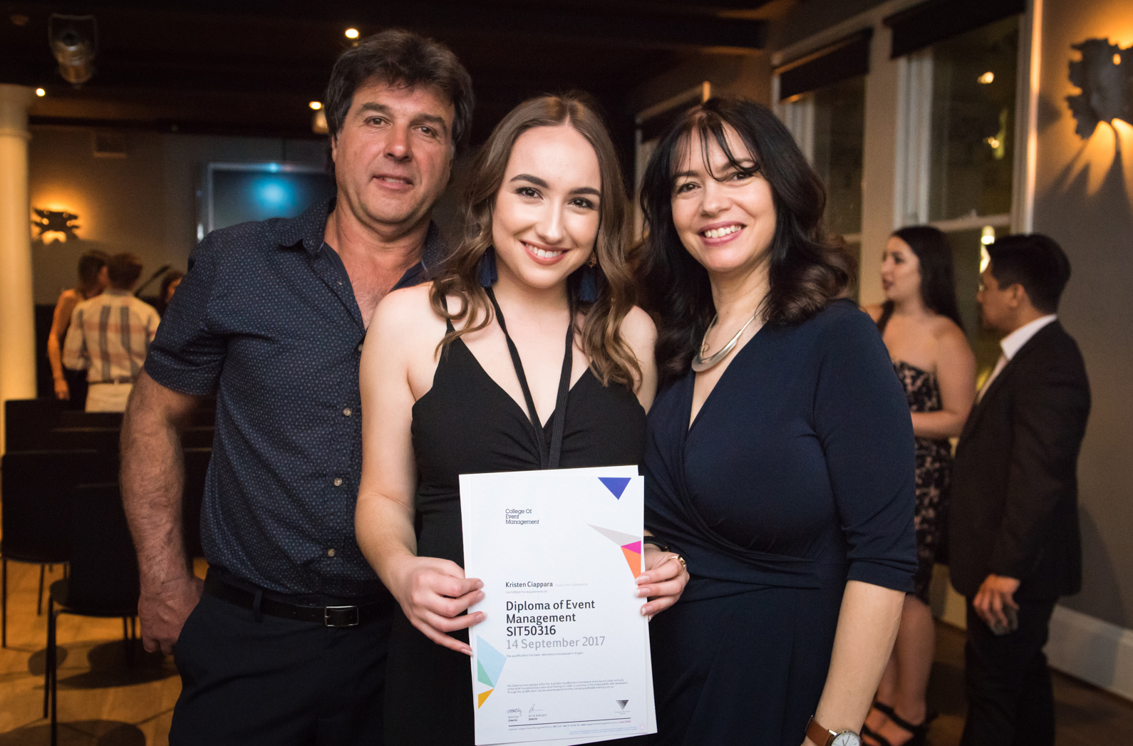 Featured graduate Kristen standing between proud parents displaying her Diploma at graduation